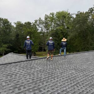 Three Coastline Roofing crew members perform a final inspection on a newly installed asphalt shingle roof surrounded by trees.
