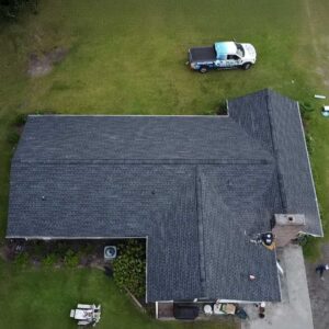 Aerial view of a completed asphalt shingle roof replacement with a Coastline Roofing truck parked nearby and a debris trailer beside the home.